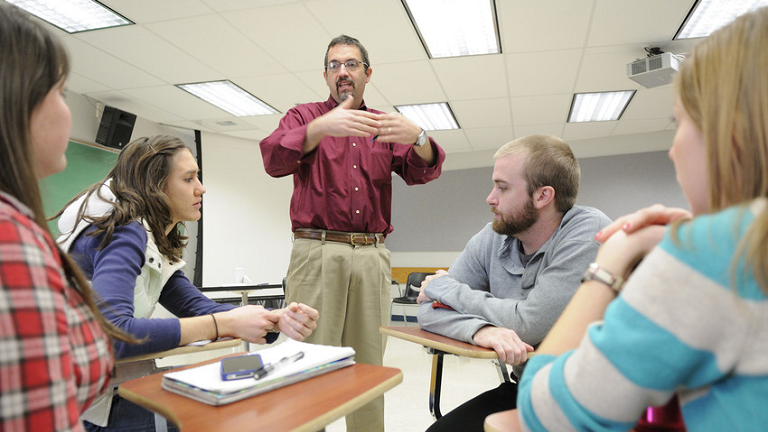 Instructor standing in classroom with four students at desks listening.