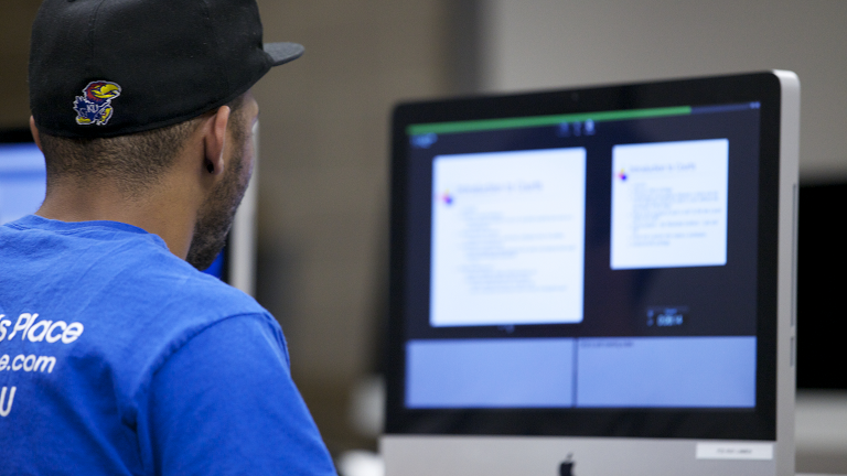 Back of student in ball cap with Jayhawk looking at a blurred out monitor screen
